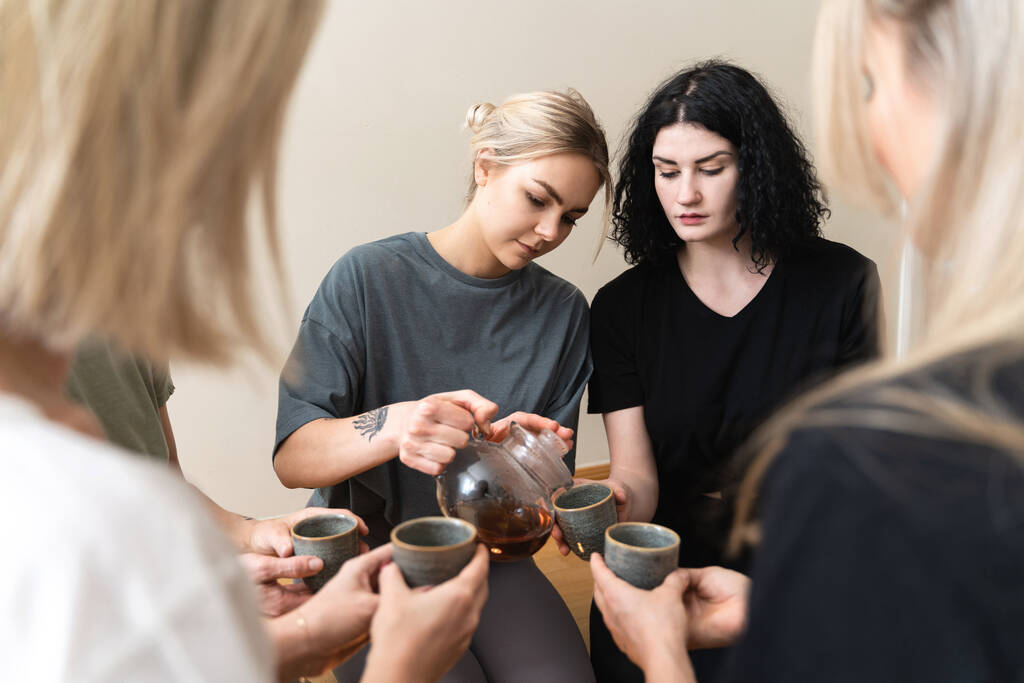 Νεαρή γυναίκα ρίχνει ζεστό τσάι στους φίλους της που κάθονται γύρω κατά τη διάρκεια μιας τελετής τσαγιού. - Φωτογραφία, εικόνα