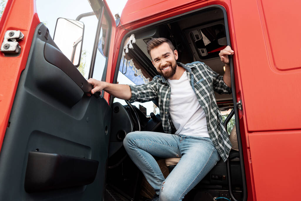 Молодой улыбчивый водитель грузовика внутри красного грузовика доволен своей работой. - Фото, изображение