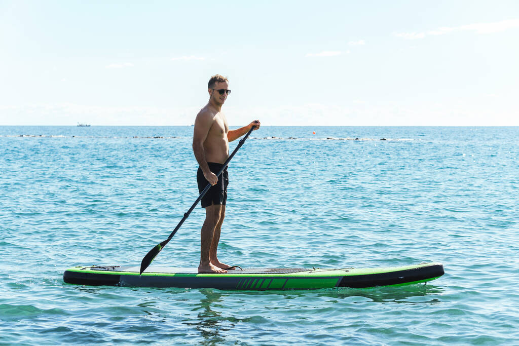 若い男性サーファーはスタンドアップパドルボードに乗っており、海の中でパドルで漕いでいます. - 写真・画像