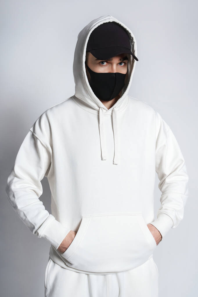 Young man wearing white hoodie, black baseball cap and cloth face mask against gray background - Photo, Image