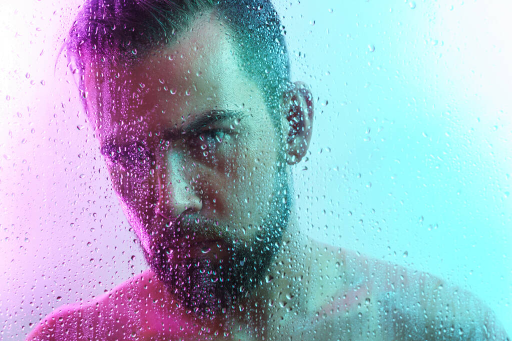 Portrét pohledného mladého muže zachyceného přes mokré sklo v barevném neonovém světle - Fotografie, Obrázek