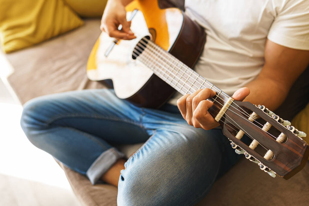 Νεαρός που κάθεται στον καναπέ παίζει άνετα ακουστική κιθάρα σε ένα φωτεινό δωμάτιο. - Φωτογραφία, εικόνα