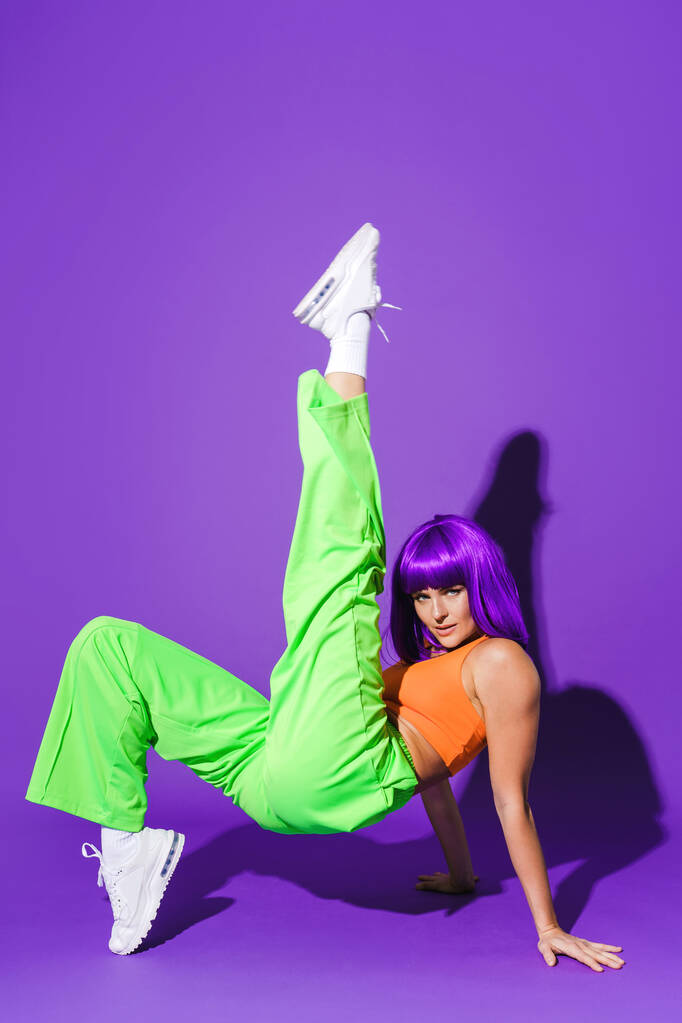 Ξέγνοιαστη χορεύτρια που φοράει πολύχρωμα αθλητικά ρούχα διασκεδάζοντας με μωβ φόντο - Φωτογραφία, εικόνα