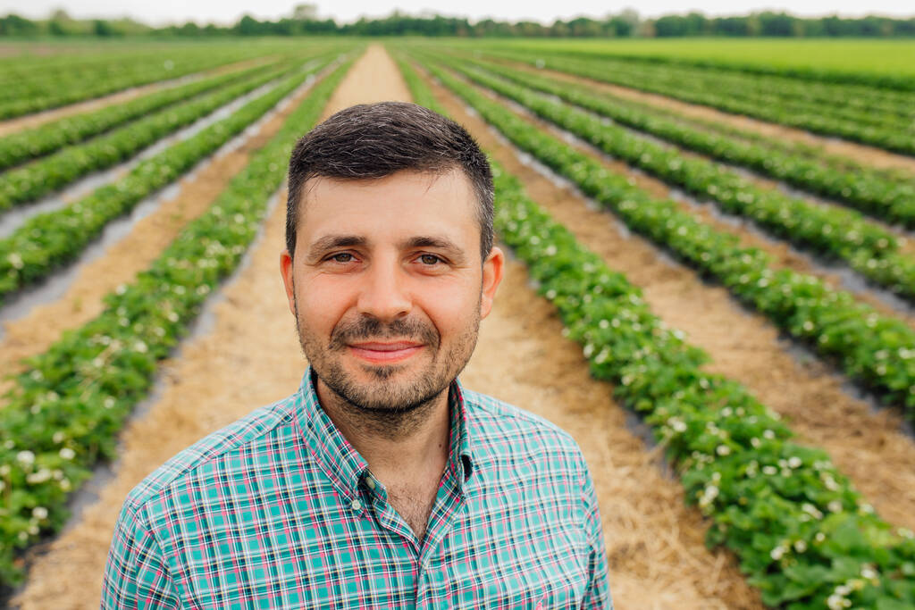 joven agricultor barbudo de nacionalidad europea mira a la cámara y sonríe. agricultor masculino vestido con camisa con cuadrados está en el campo agrícola - Foto, imagen