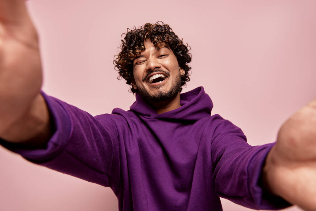 Low angle view of happy young Indian man making selfie against pink background - Photo, Image