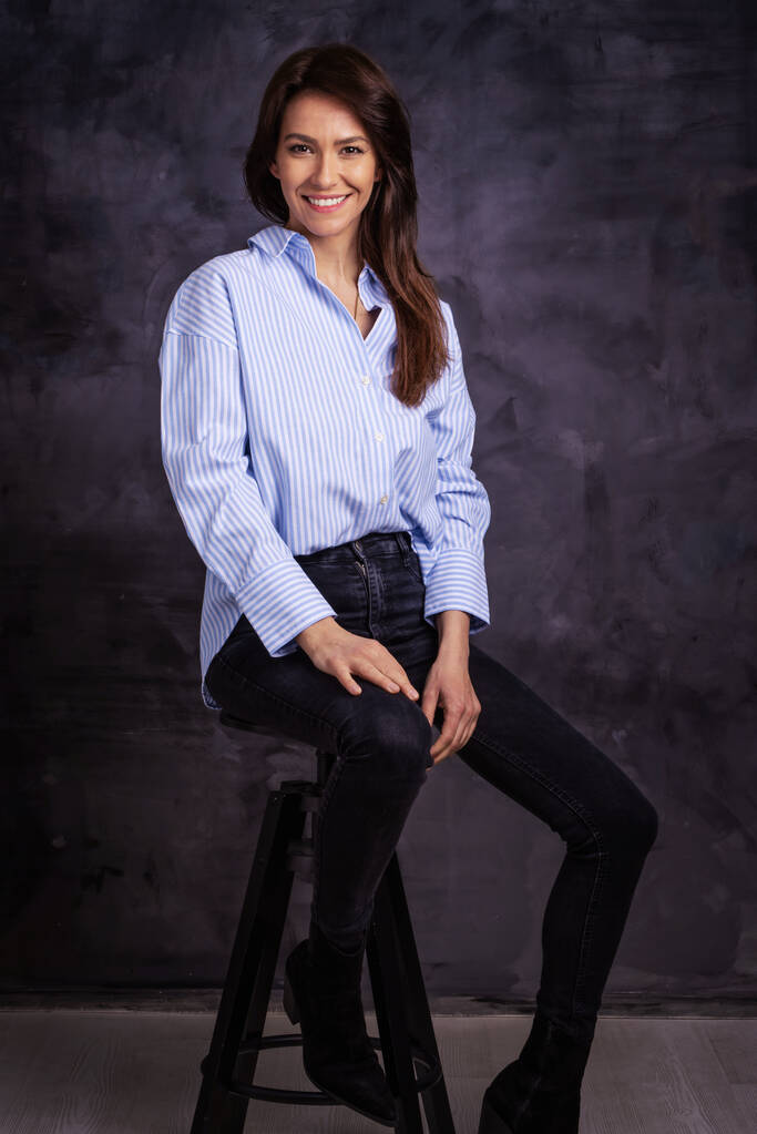 Estúdio completo tiro de uma mulher atraente vestindo camisa e jeans enquanto sentado em fundo escuro isolado.. Espaço de cópia.  - Foto, Imagem