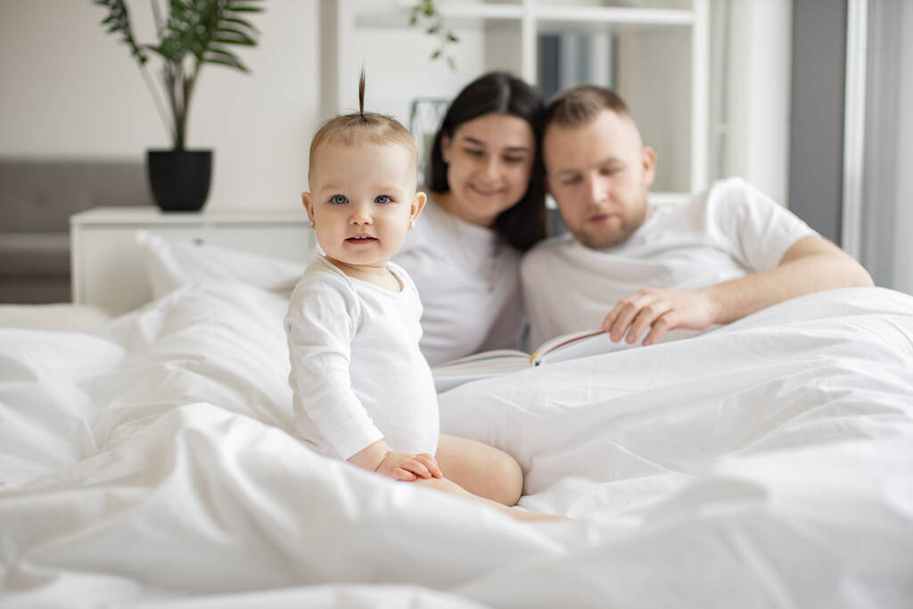 Cheerful little kid in white baby clothes resting on bed while joyful adult people staying under blanket in bright bedroom interior. Loving mother and father telling nursery rhyme from verse book. - Photo, Image