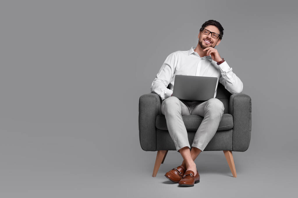 Hombre feliz con portátil sentado en sillón sobre fondo gris. Espacio para texto - Foto, imagen