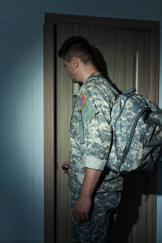 zijaanzicht van Amerikaanse militaire man met rugzak staan in de buurt ingang deur thuis - Foto, afbeelding