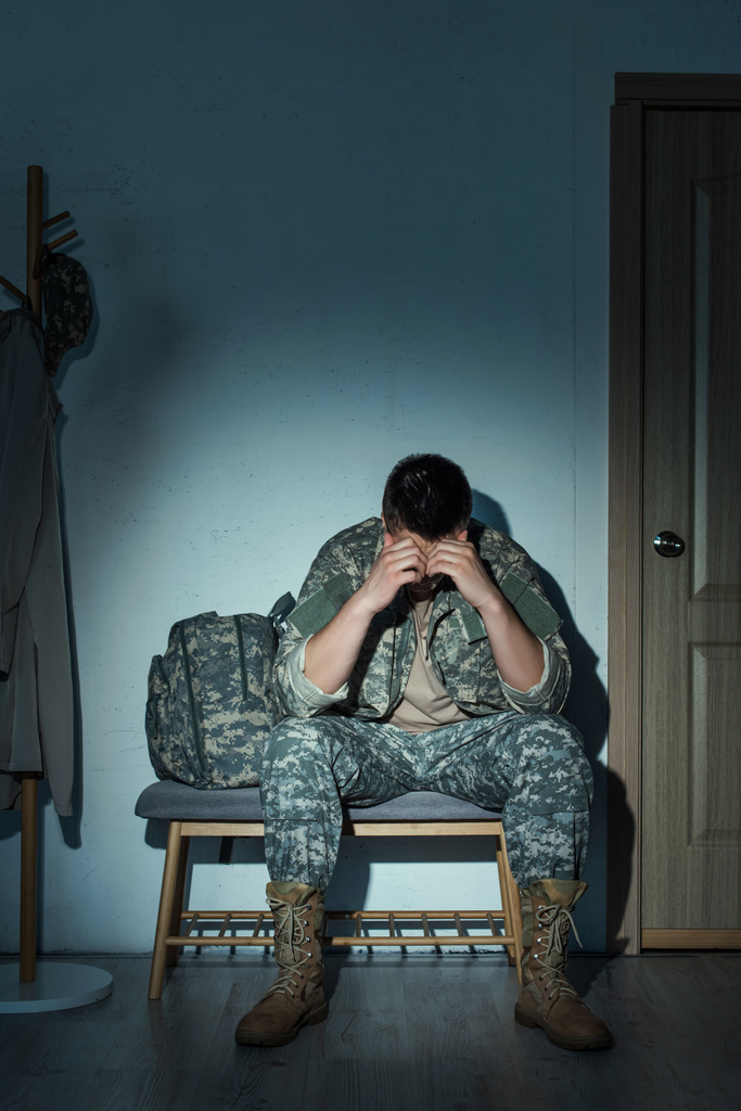 Soldado en uniforme que sufre de trastorno de estrés postraumático mientras está sentado en el pasillo en casa por la noche  - Foto, imagen