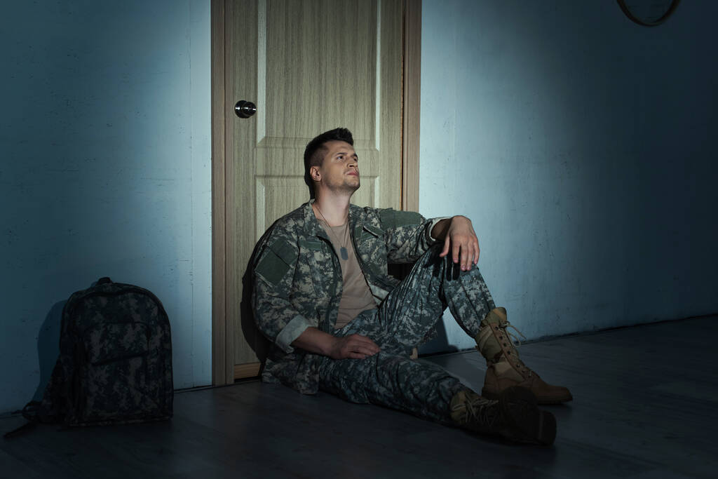 バックパックと廊下のドアの近くの床の上に夜に座って落ち込んで兵士  - 写真・画像