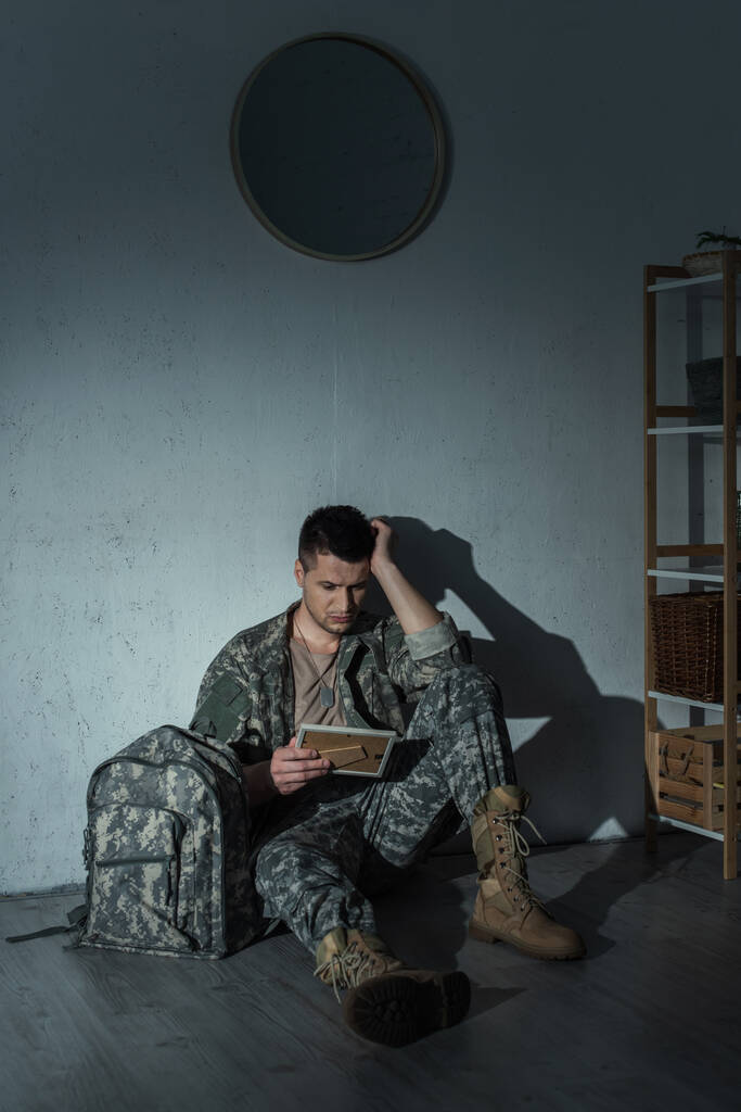 Przygnębiony żołnierz trzymający ramę ze zdjęciem siedząc w pobliżu plecaka w domu w nocy  - Zdjęcie, obraz