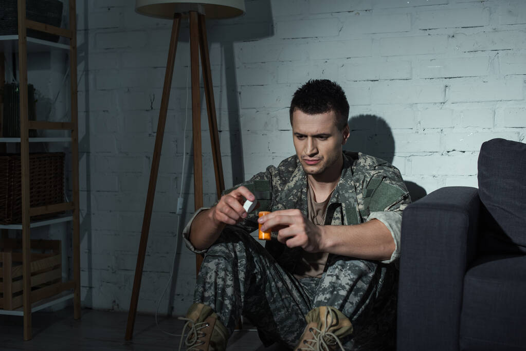 Soldat in Uniform mit Medikamenten, während er nachts zu Hause an posttraumatischer Belastungsstörung leidet  - Foto, Bild