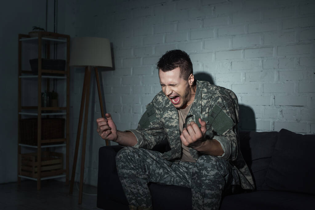 Soldat en colère avec trouble de stress post-traumatique criant sur le canapé à la maison la nuit  - Photo, image