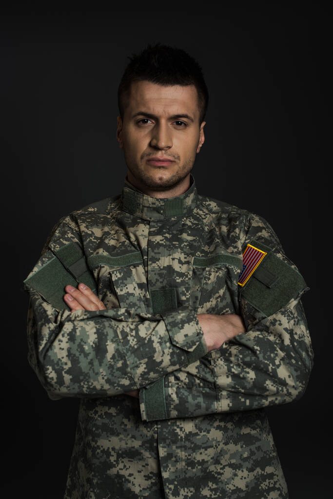 καταθλιπτικός Αμερικανός στρατιώτης με στολή που στέκεται με σταυρωμένα χέρια απομονωμένος στα μαύρα  - Φωτογραφία, εικόνα