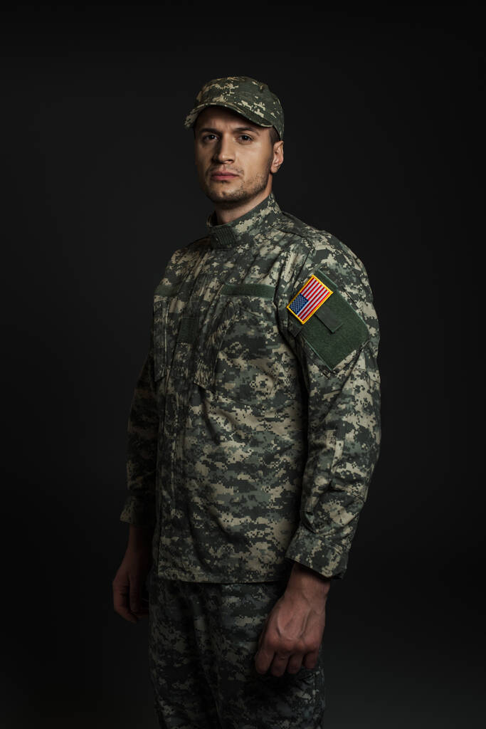 patriotyczny amerykański żołnierz w mundurze i czapce patrząc na aparat odizolowany na czarny  - Zdjęcie, obraz