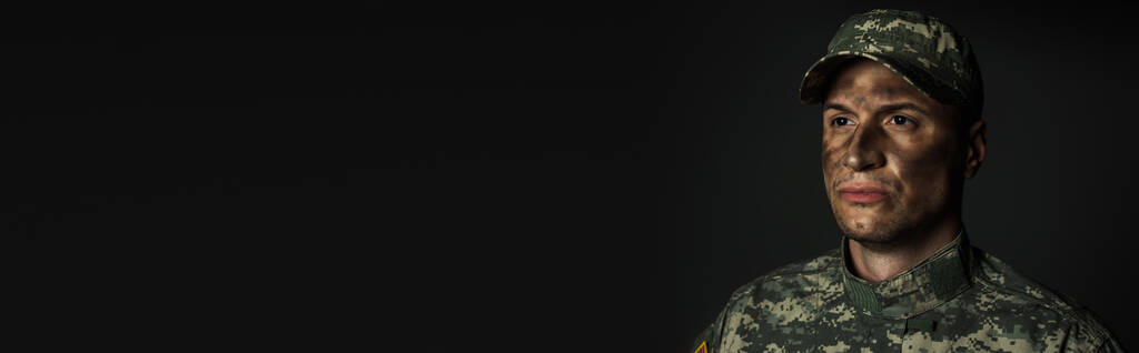 военный с грязью на лице стоя в униформе и шапке изолированы на сером, баннер   - Фото, изображение