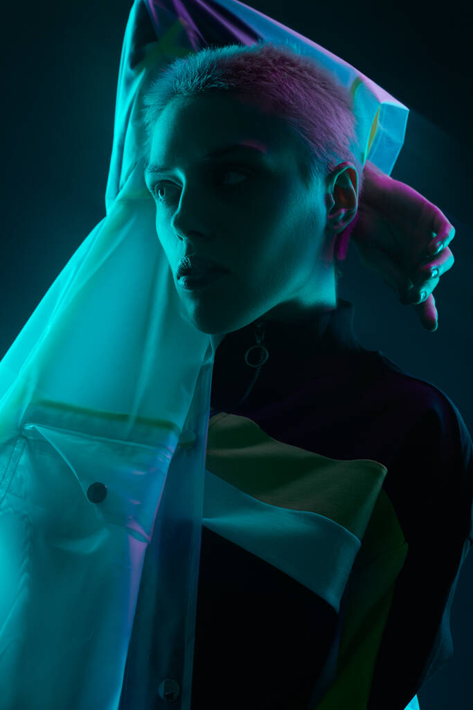 Serieuze jonge vrouw model met shirt blond haar in transparante regenjas weg te kijken aandachtig in donkere studio met blauwe neon verlichting - Foto, afbeelding