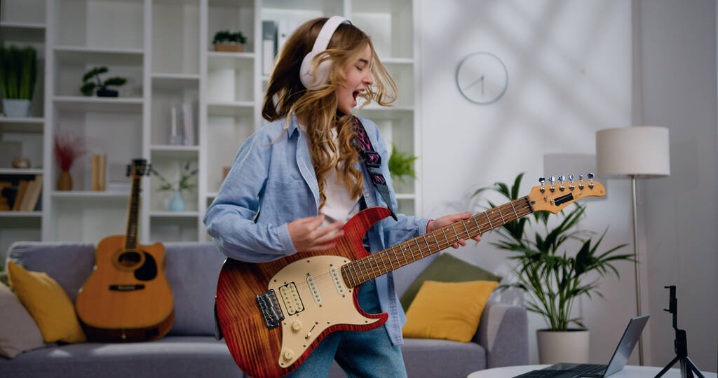Uzaktan kumandalı beyaz kulaklık takan neşeli genç kız evde elektro gitar çalıyor ve modern müzikten zevk alıyor. Müzik aşığı kavramı. - Fotoğraf, Görsel