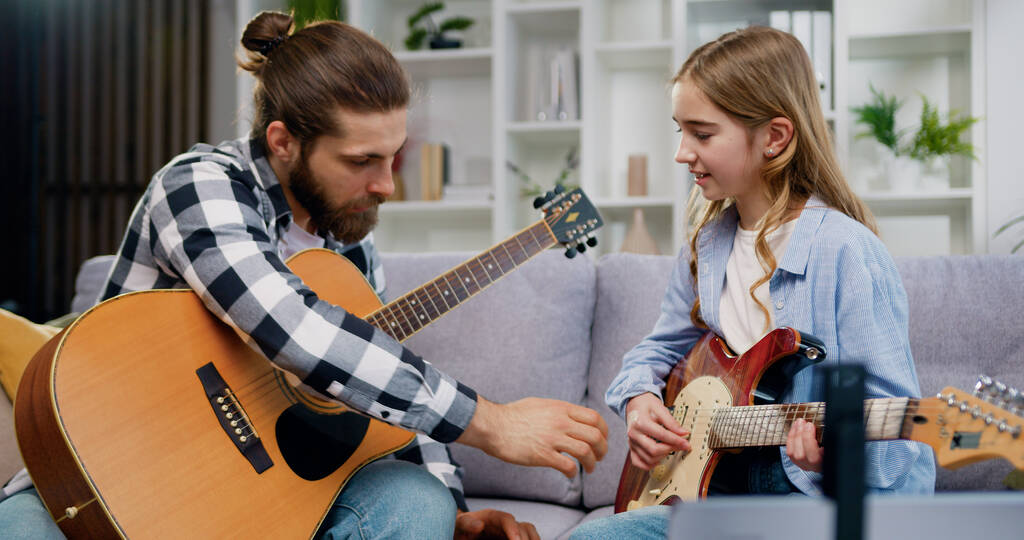 Pretty caucasian 10 year old girl practicing playing electric guitar and her father teaching her, spending leisure time together during a music lesson while sitting on the sofa at home. - Photo, Image