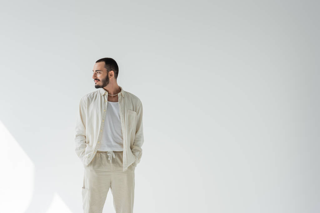 Smiling homosexual man in beige clothes and pearl necklace holding hands in pockets of pants and looking away while standing on grey background with sunlight  - Photo, Image