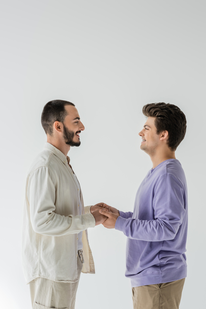 Sivunäkymä hymyilevästä homoseksuaalisesta parista rennoissa vaatteissa, jotka katsovat toisiaan ja pitävät toisiaan kädestä seistessään harmaalla eristyksissä  - Valokuva, kuva