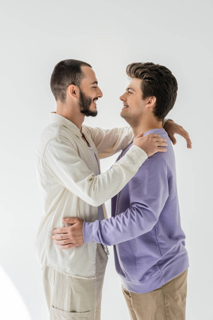 Vista lateral de pareja homosexual joven y alegre en ropa casual abrazándose y mirándose mientras están de pie sobre un fondo gris - Foto, Imagen