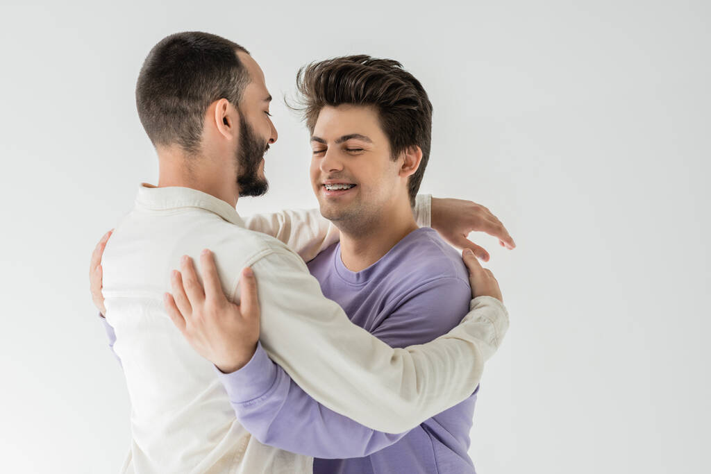 Überglückliches brünettes gleichgeschlechtliches Paar in lässiger Kleidung, das sich umarmt und die Augen schließt, während es isoliert auf grau steht  - Foto, Bild