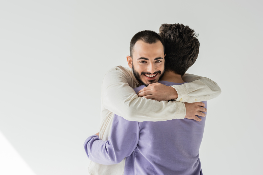 Cheerful and bearded gay man in casual clothes hugging young brunette boyfriend and looking at camera on grey background with sunlight - Photo, Image