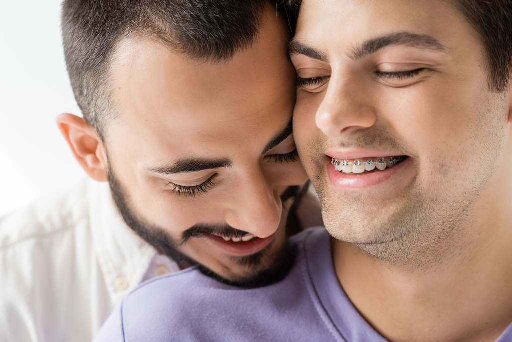 Nahaufnahme eines lächelnden und bärtigen homosexuellen Mannes, der in der Nähe seines jungen Partners steht, mit geschlossenen Augen und Zahnspangen an Zähnen isoliert auf grau  - Foto, Bild
