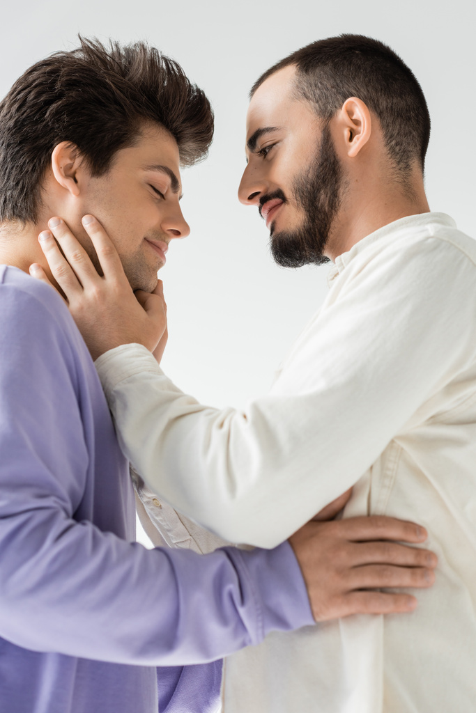 Vista lateral do homem homossexual barbudo despreocupado em roupas casuais tocando o rosto do namorado com os olhos fechados enquanto estão juntos isolados em cinza  - Foto, Imagem