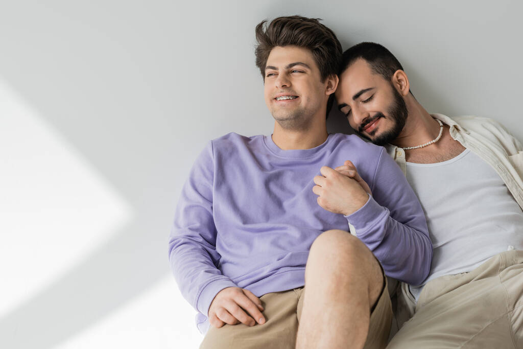 Souriant jeune homme gay en vêtements décontractés tenant la main du petit ami barbu avec les yeux fermés tout en étant assis ensemble sur fond gris avec la lumière du soleil  - Photo, image