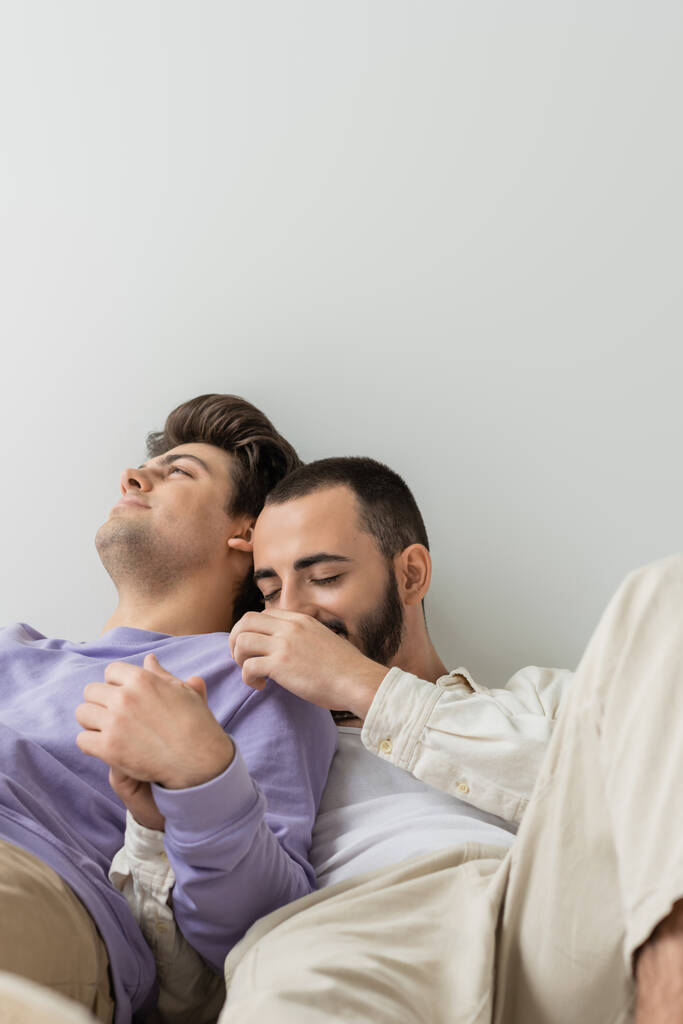 Young homosexual man touching shoulder and holding hand of brunette boyfriend in casual clothes while sitting together on grey background - Photo, Image