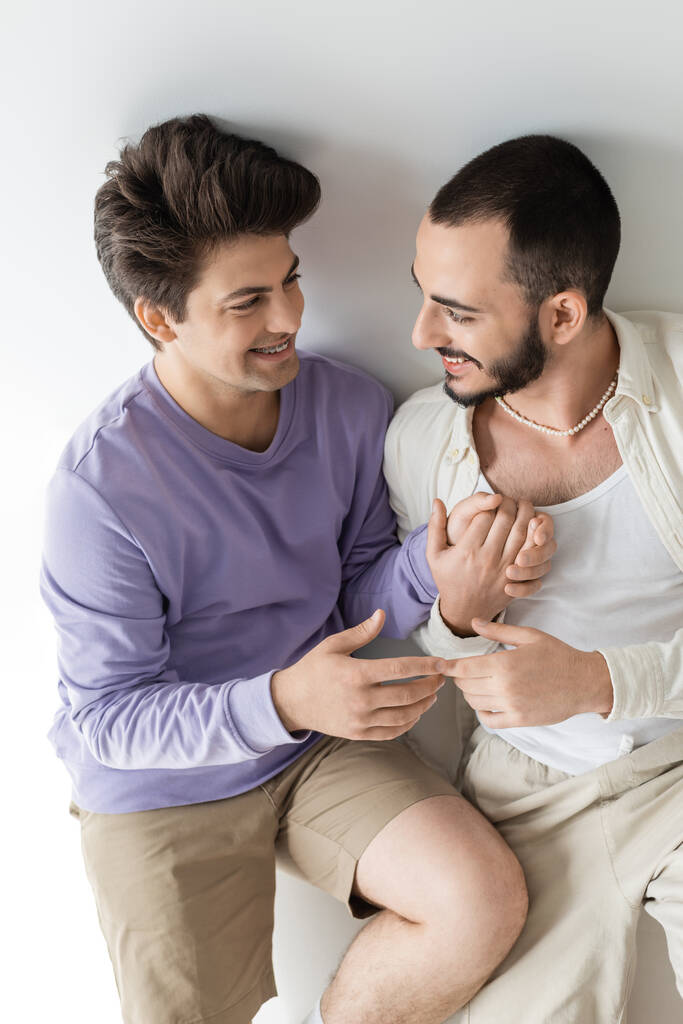 Magas látószögű kilátás fiatal és vidám homoszexuális pár alkalmi ruhák fogják egymás kezét, miközben beszél, és együtt ülnek szürke háttér - Fotó, kép