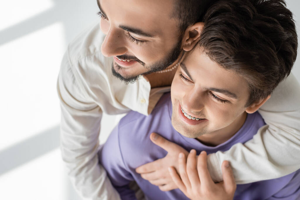 Overhead view of positive and bearded homosexual man hugging young boyfriend in purple sweatshirt and braces on grey background with sunlight  - Photo, Image