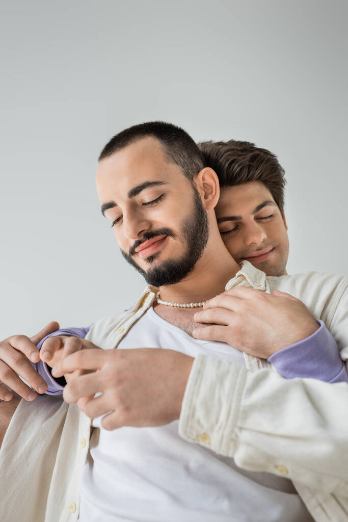 Portret uśmiechniętego i brodatego homoseksualisty w luźnych ciuchach i perłowym naszyjniku dotykającym dłoni brunetki chłopaka zamykającego oczy na szarości  - Zdjęcie, obraz