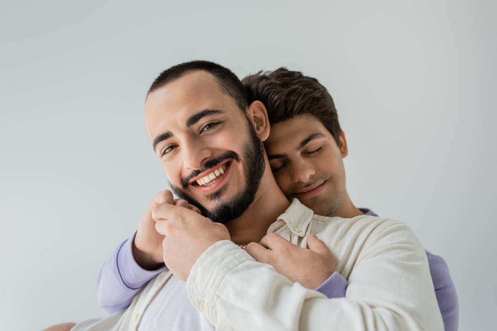 Портрет позитивного и бородатого гея, держащегося за руку молодого парня-брюнетки возле лица и смотрящего на камеру, изолированную на сером  - Фото, изображение