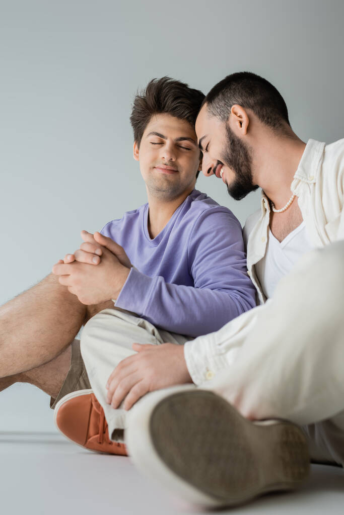 jong en positief hetzelfde geslacht paar met gesloten ogen in casual kleding houden handen terwijl zitten en rusten samen op grijze achtergrond - Foto, afbeelding