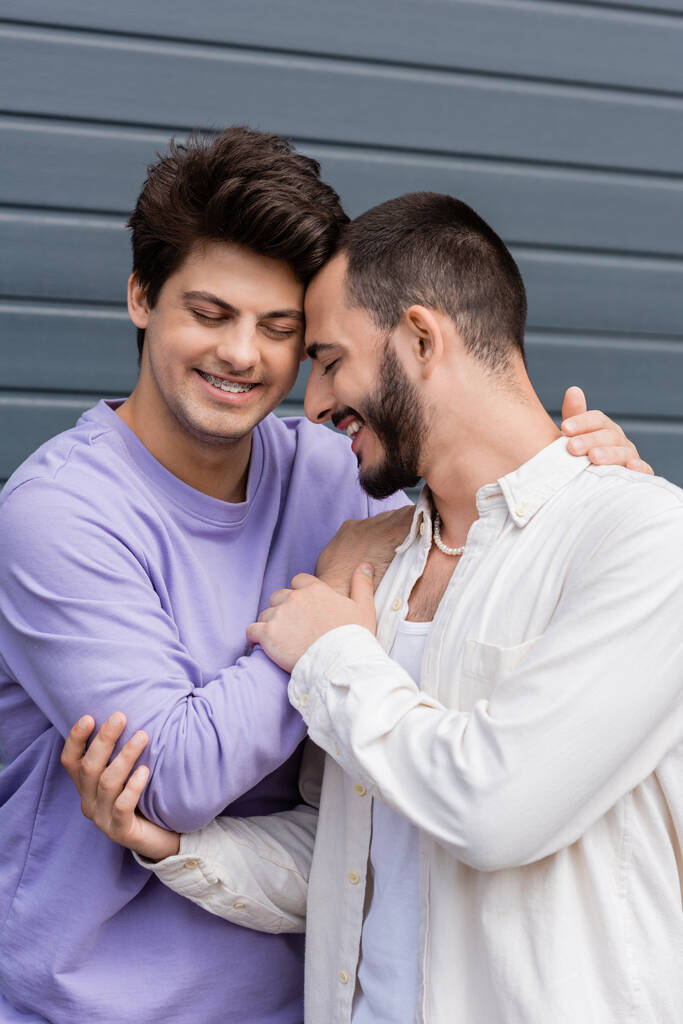 Πορτρέτο του θετικού ομοφυλόφιλου άνδρα σε casual ρούχα και τιράντες αγκαλιάζει γενειοφόρος φίλος σε πουκάμισο και μαργαριτάρι κολιέ με κλειστά μάτια, ενώ στέκεται κοντά σε κτίριο σε εξωτερικούς χώρους  - Φωτογραφία, εικόνα