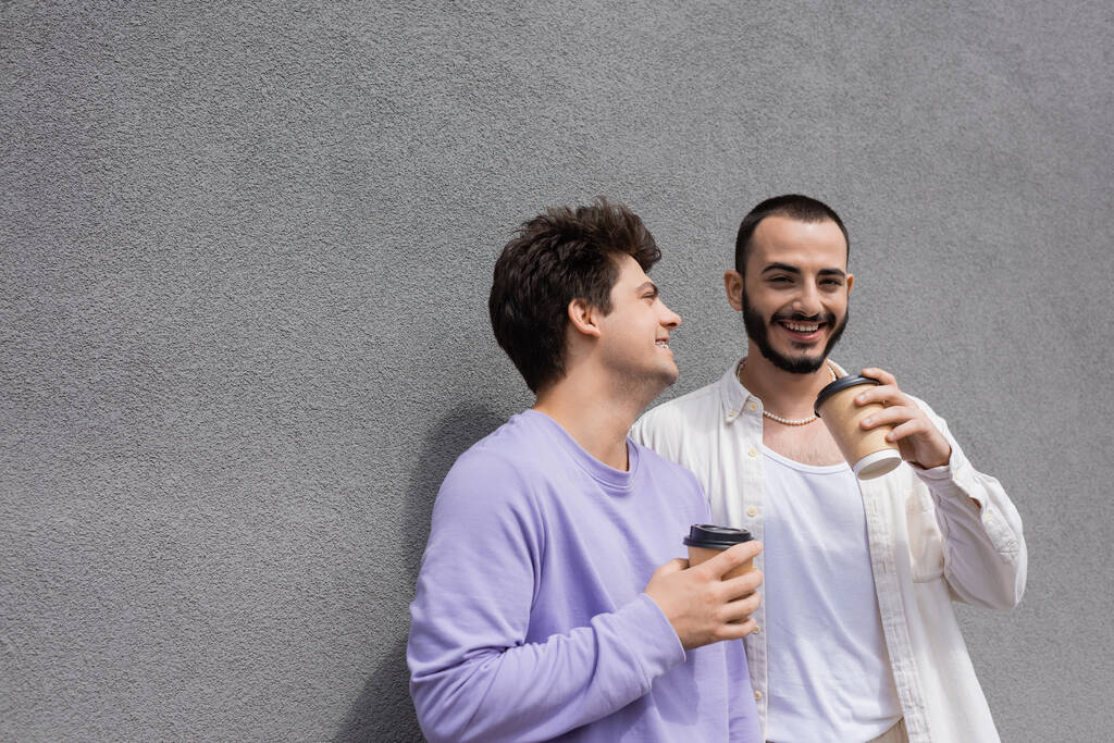 Felice uomo omosessuale barbuto in possesso di caffè per andare vicino fidanzato in felpa viola e bretelle mentre in piedi vicino edificio sulla strada urbana  - Foto, immagini