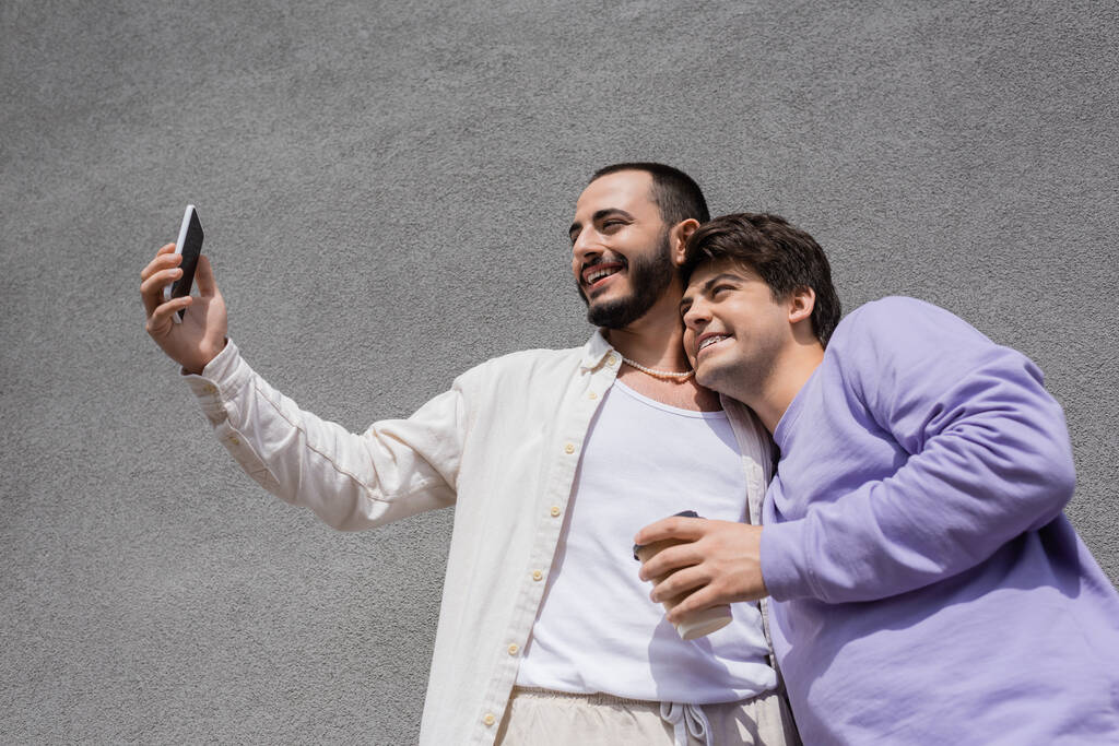 Nízký úhel pohledu na pozitivní a vousatý gay muž, přičemž selfie s bezstarostným přítelem držící odnášející kávu v blízkosti budovy venku ve dne  - Fotografie, Obrázek