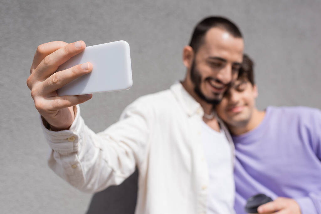 Blurred same sex couple in casual clothes taking selfie on smartphone together while standing near building wall on urban street at daytime - Photo, Image