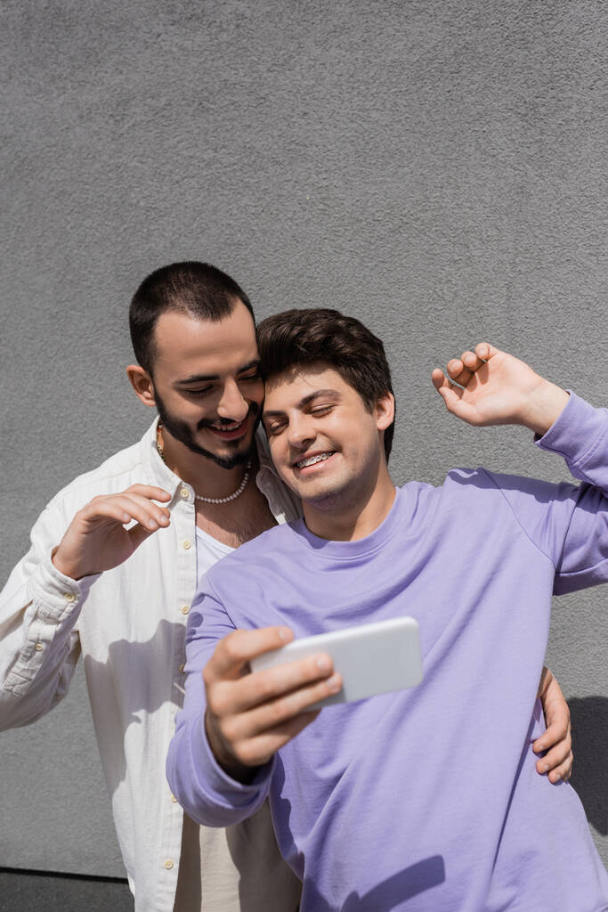 Iloinen homoseksuaalinen mies rennoissa vaatteissa matkapuhelimen avulla seistessään poikaystävän vieressä hammasraudoissa ja rakennettaessa kaupunkien kadulla ulkona  - Valokuva, kuva