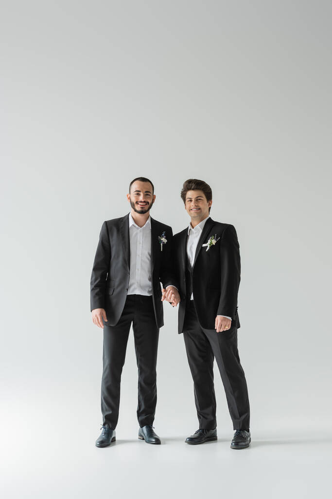 Pełna długość młodych i uśmiechniętych homoseksualistów w klasycznych garniturach z butonierkami trzymającymi się za ręce i patrzącymi w kamerę na szarym tle  - Zdjęcie, obraz