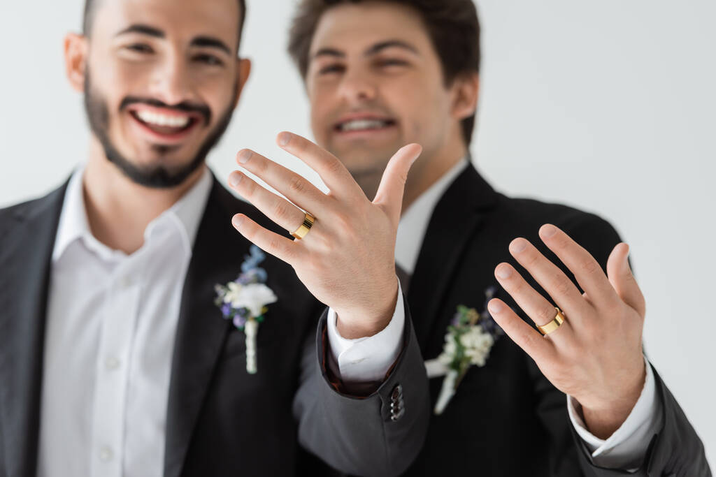 Burred giovani sposi omosessuali in abiti classici che mostrano anelli nuziali dorati alla macchina fotografica durante la celebrazione del matrimonio isolati su grigio   - Foto, immagini