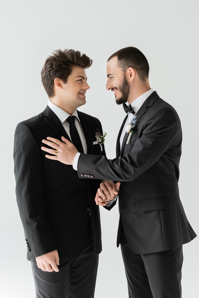 Веселый и бородатый гей в элегантной формальной одежде с цветочным бутоньером, держащимся за руку жениха во время свадебного торжества, изолированного на сером   - Фото, изображение