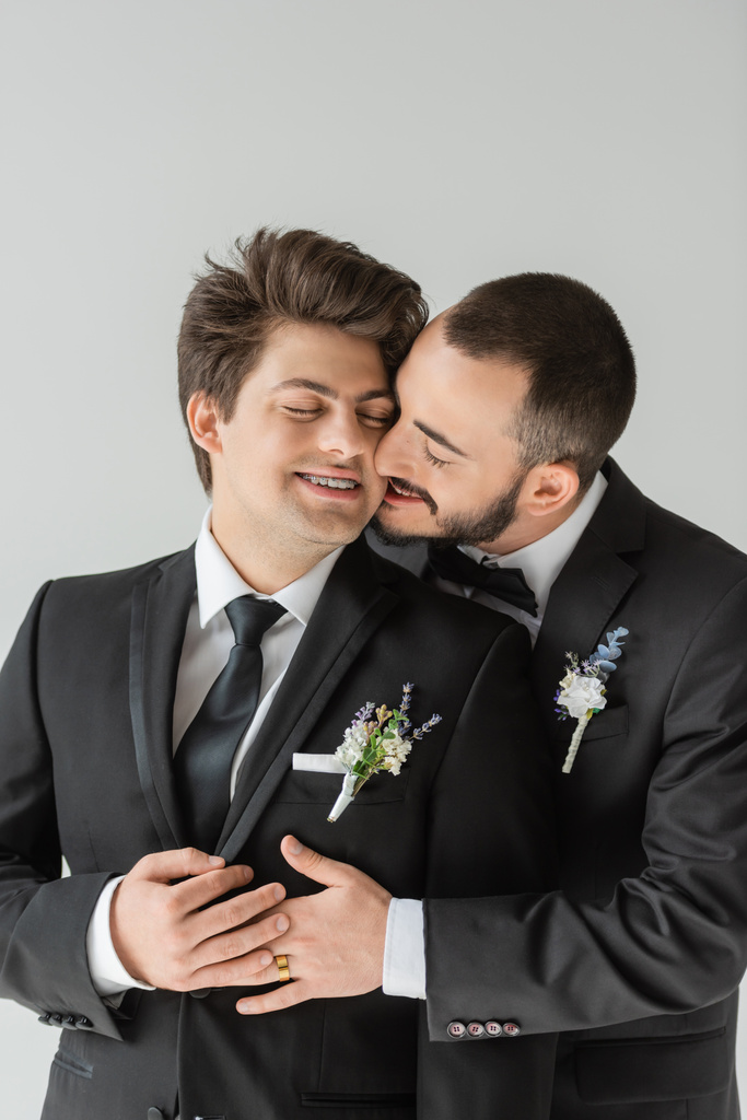 Giovane e barbuto sposo gay in abito elegante con boutonniere abbracciare e baciare fidanzato sorridente con gli occhi chiusi e bretelle mentre celebra il matrimonio isolato su grigio   - Foto, immagini