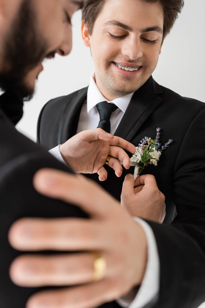 niewyraźne i brodaty gej pan młody dostosowując kwiatowy boutonniere na elegancki garnitur młody chłopak z aparat ortodontyczny podczas ślub uroczystości odizolowany na szary   - Zdjęcie, obraz