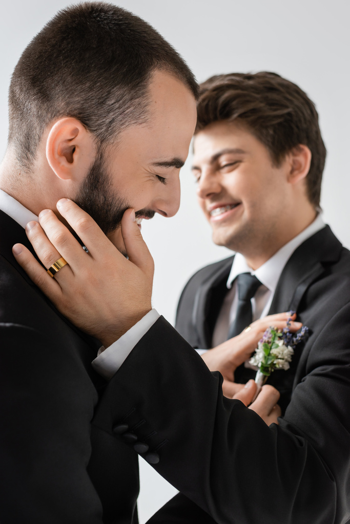 Veselý vousatý gayové ženich nastavení rozmazané květinové boutonniere na oblek rozmazané usmívající se mladý přítel v rovnátka během svatební oslavy izolované na šedé   - Fotografie, Obrázek
