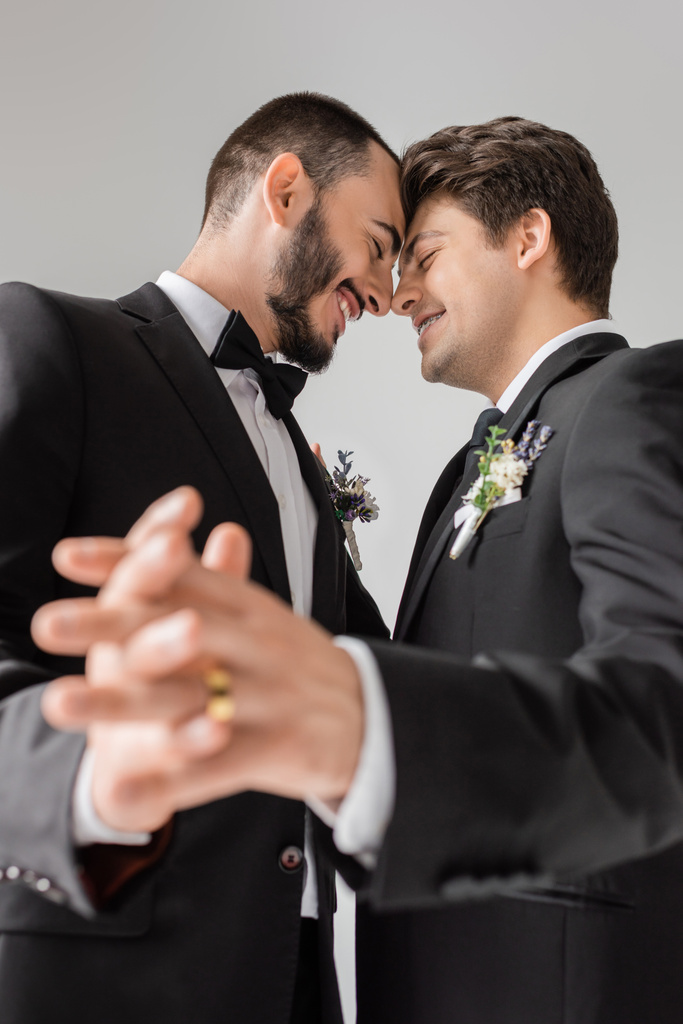 Low angle view of cheerful gay couple in formal wear with floral boutonnieres holding blurred hands during wedding isolated on grey  - Photo, Image